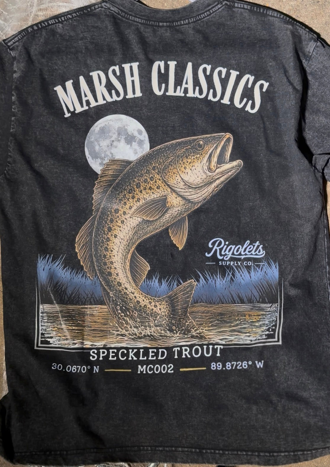 Speckled trout fishing shirt, black vintage washed with moonlit speckled trout jumping out of the water. With the Rigolets Supply logo on it 