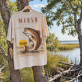 A beige graphic t-shirt hanging on a wooden hanger from a tree near a lake, featuring a large bass fish illustration and the text 'MARSH' and 'Florida'.